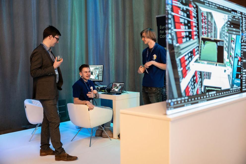 Three men having a conversation in the Demo Lab at EuroHPC Summit 2023. One is sitting at a computer and there is a translucent screen with lines of code in the foreground.